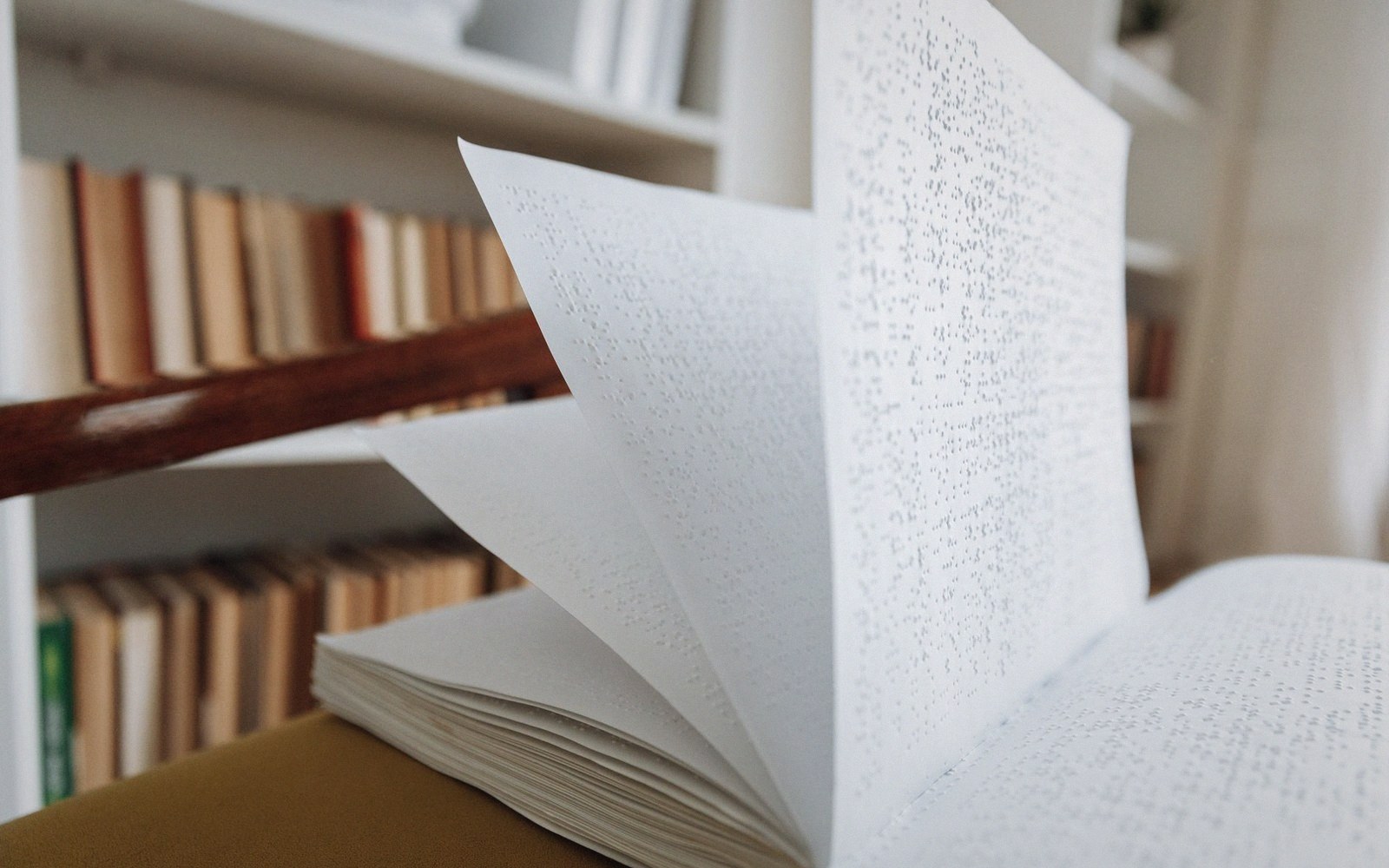An open braille book with pages fanned out on a shelf beside bound volumes