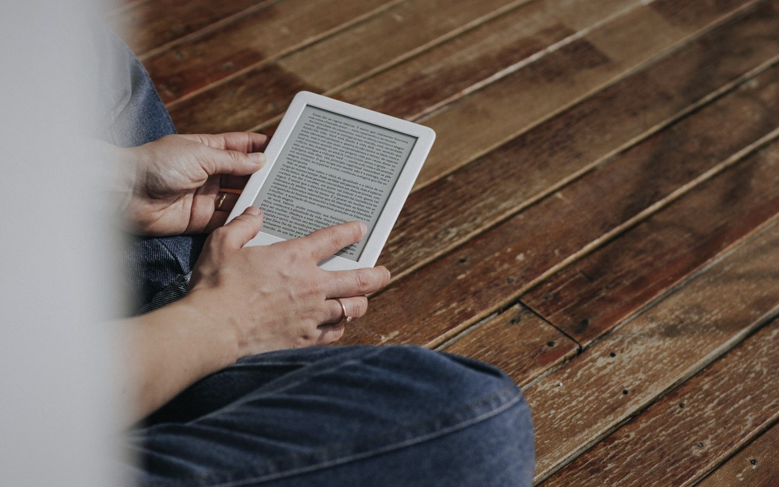 Person seated on a wooden floor reading text on a white e-reader device