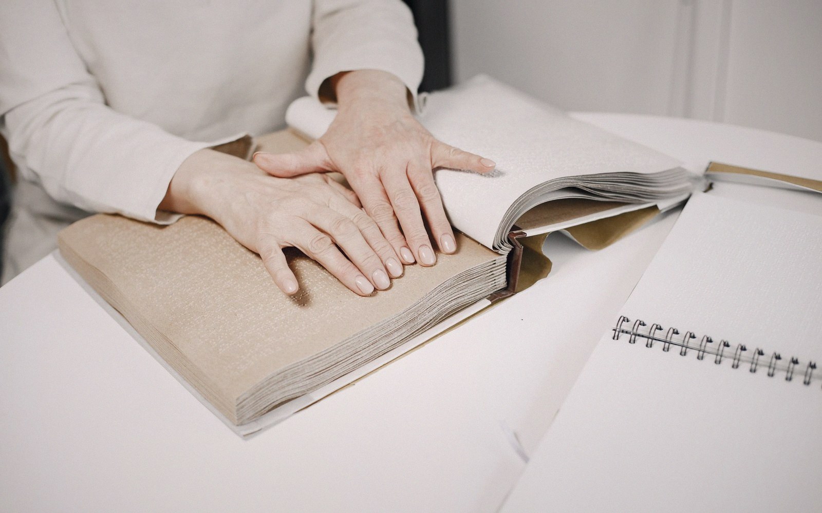 Hands resting on an open book with thick, textured pages beside a spiral notebook