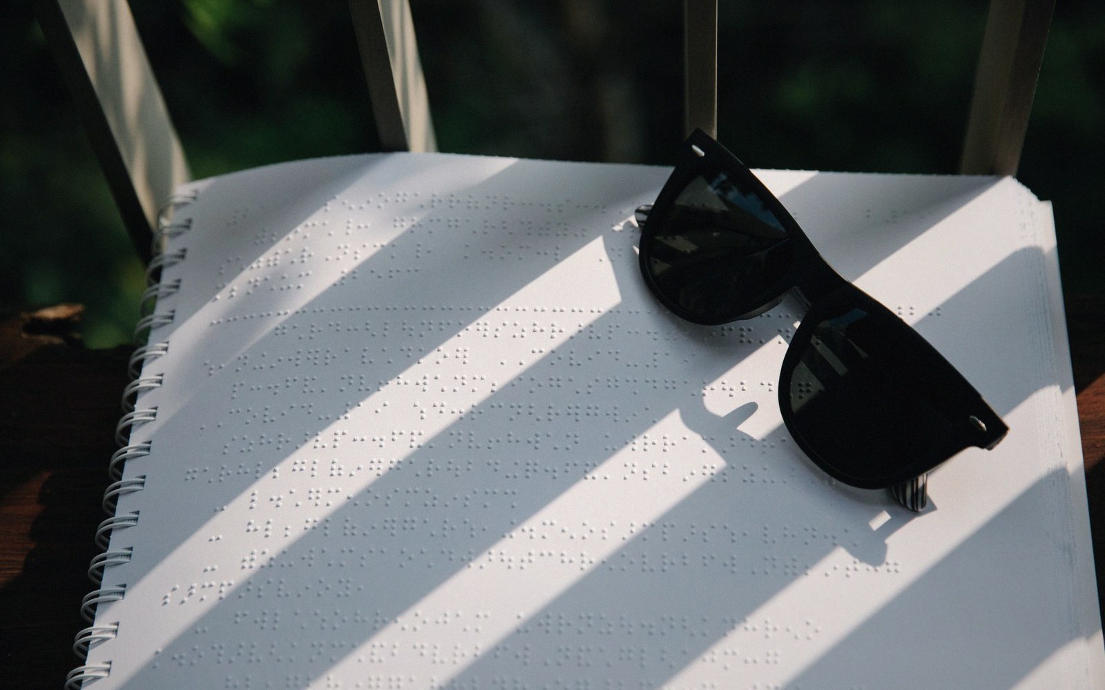 Sunglasses resting on a spiral-bound braille notebook with sunlight casting diagonal shadows